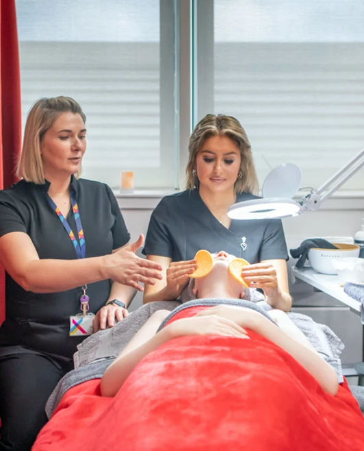 A lecturer demonstrates a facial massage technique to a beauty therapy student who is performing a treatment on a mannequin in a spa-like environment. A lecturer demonstrates a facial massage technique to a beauty therapy student who is performing a treatment on a mannequin in a spa-like environment.